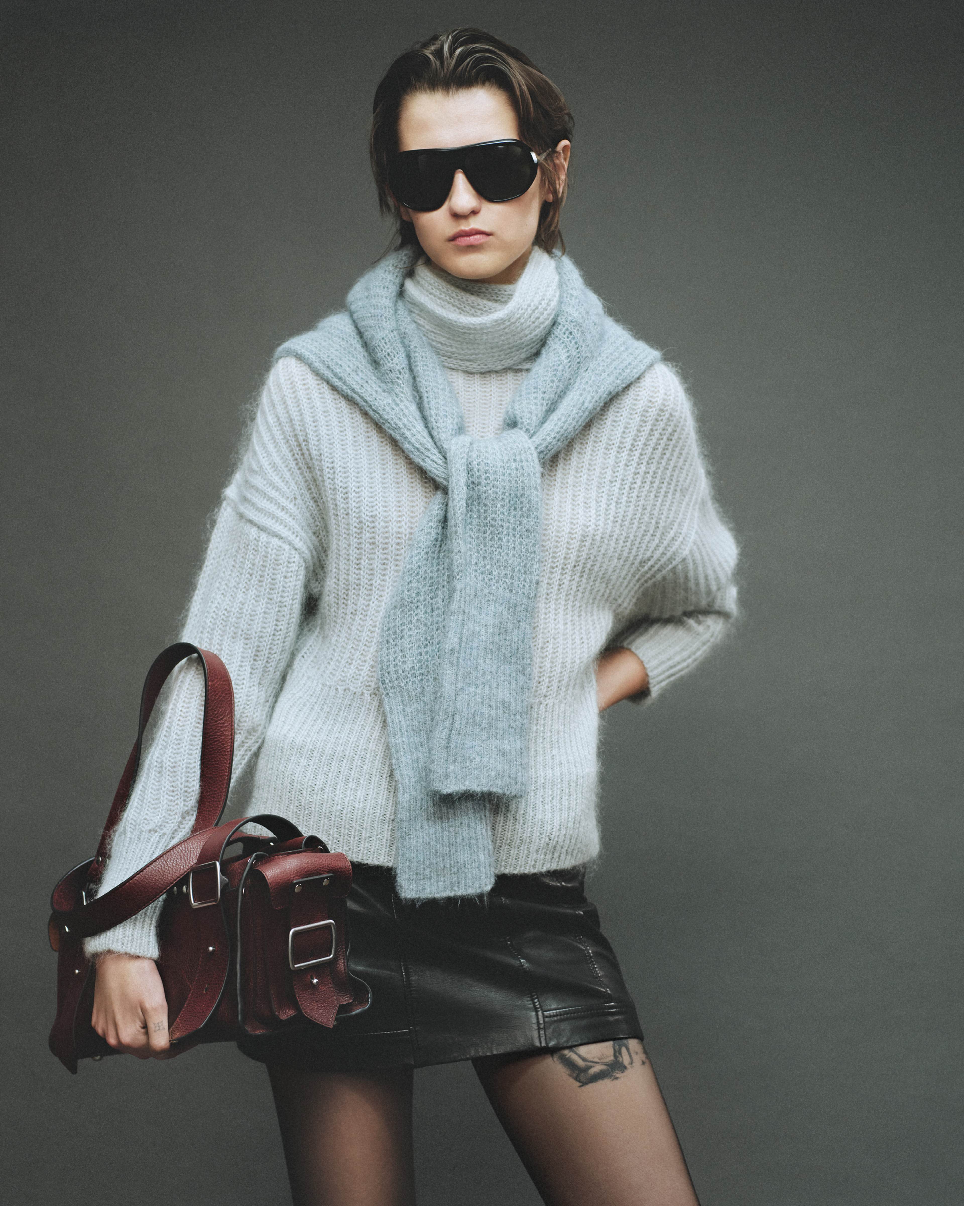 A woman with short black hair stands against a grey background wearing a black leather skirt, a light blue knitted jumper, and holding an oxblood red leather handbag.