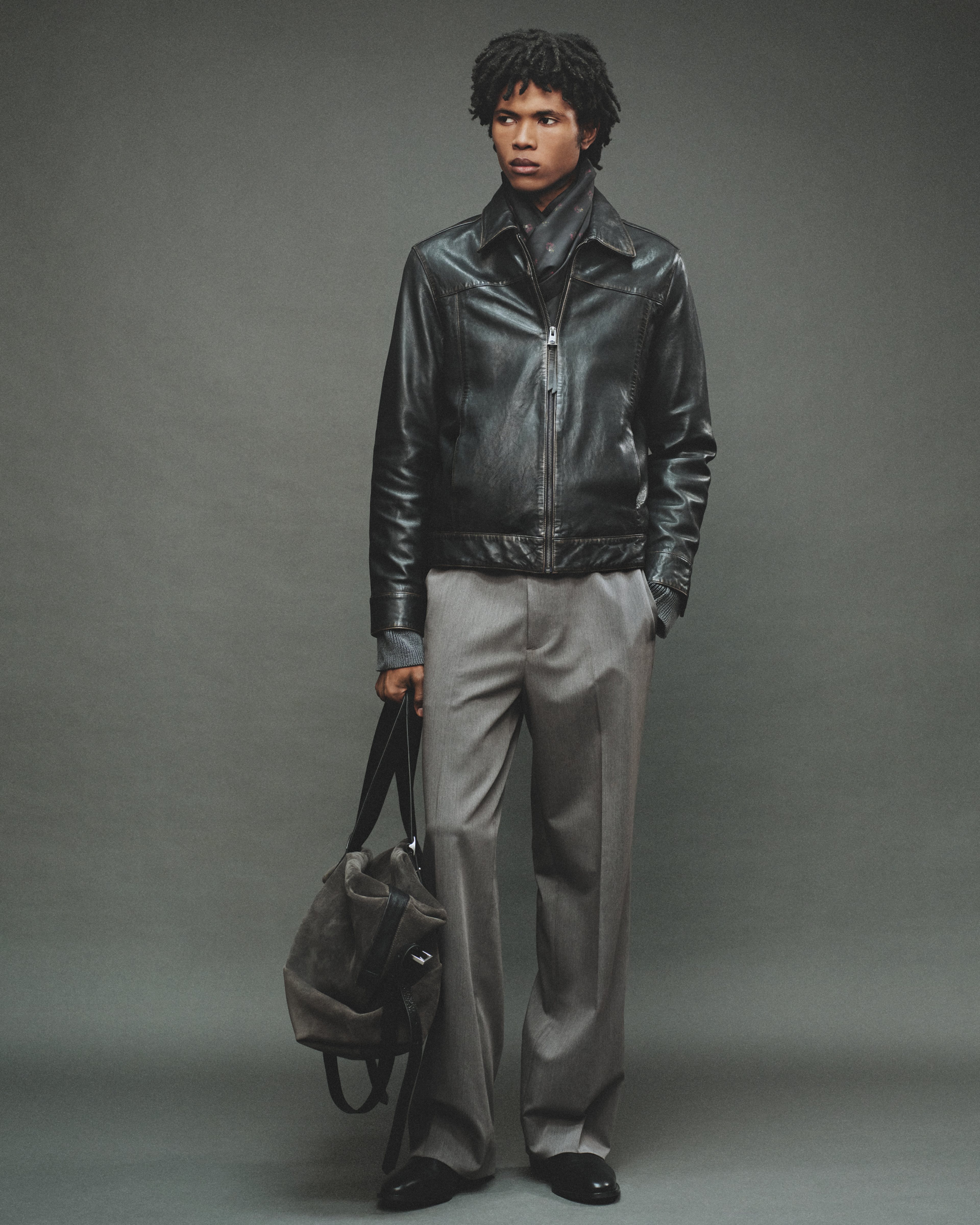 A man wearing a black leather jacket, grey tailored trousers and a suede handbag looking to the side.