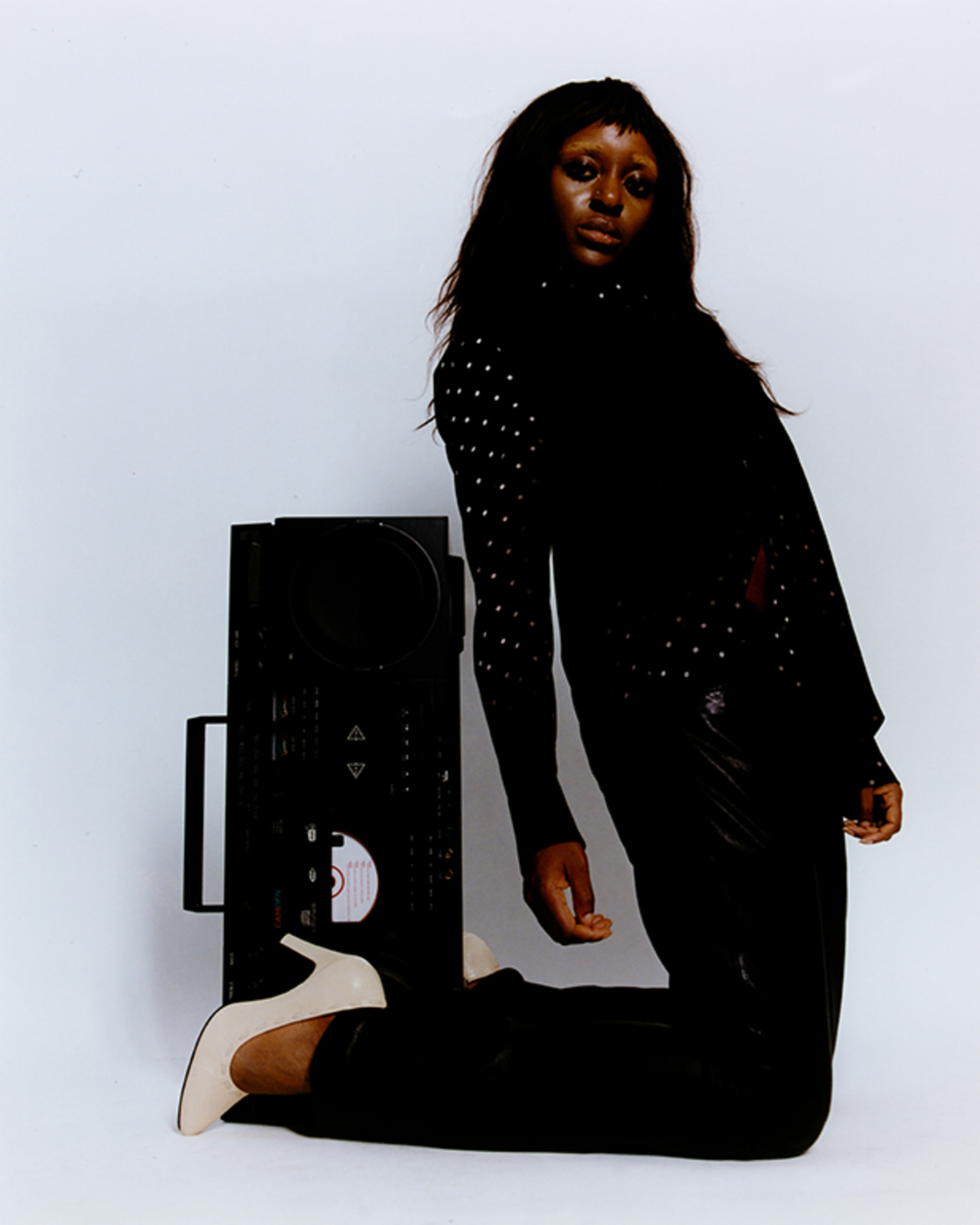 A woman in a black, studded outfit kneels beside a large speaker against a plain white backdrop, striking a calm, confident pose.
