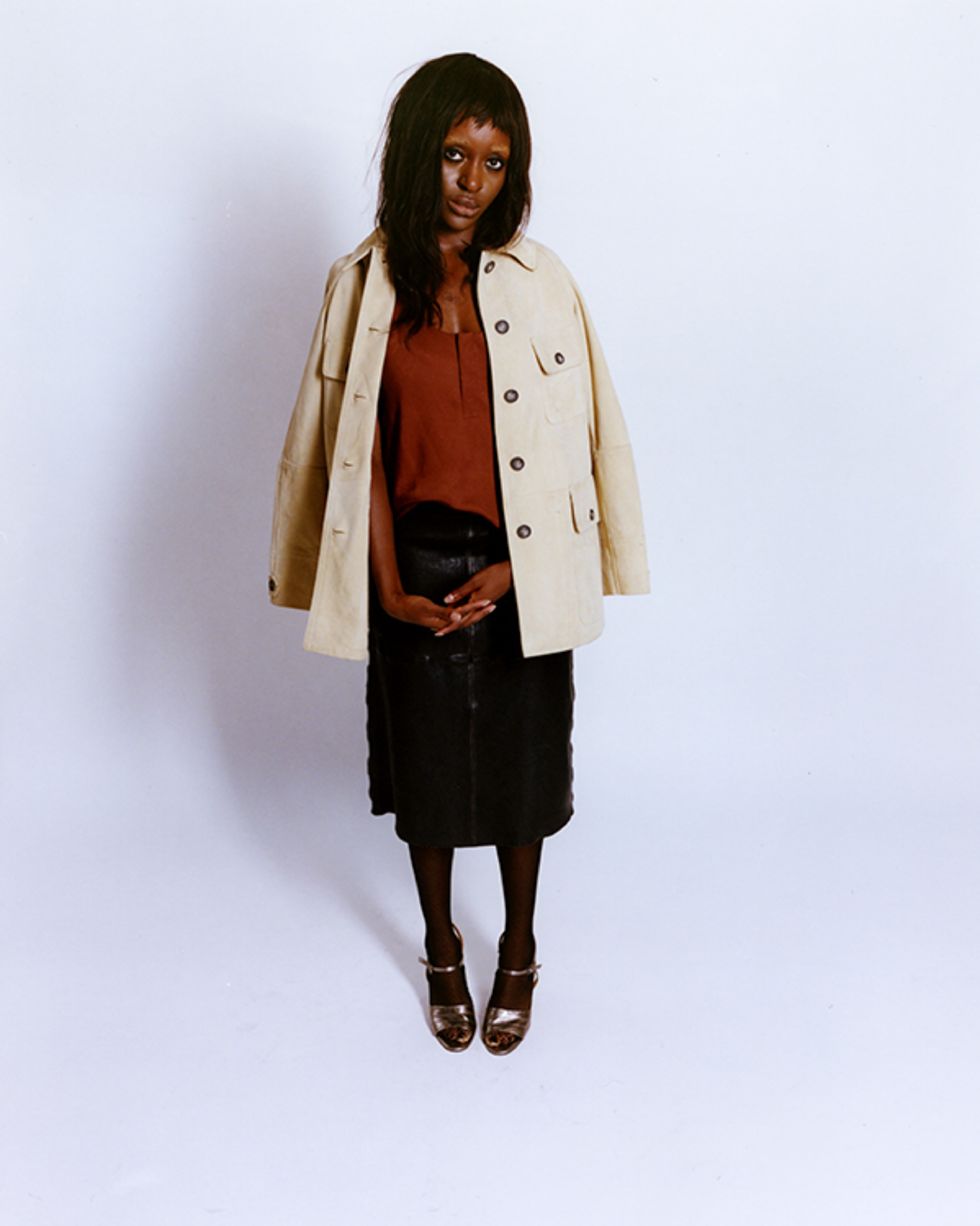 A woman stands against a pale studio backdrop in a cream coat, rust blouse, black skirt, and heels, posing with a calm, confident expression.