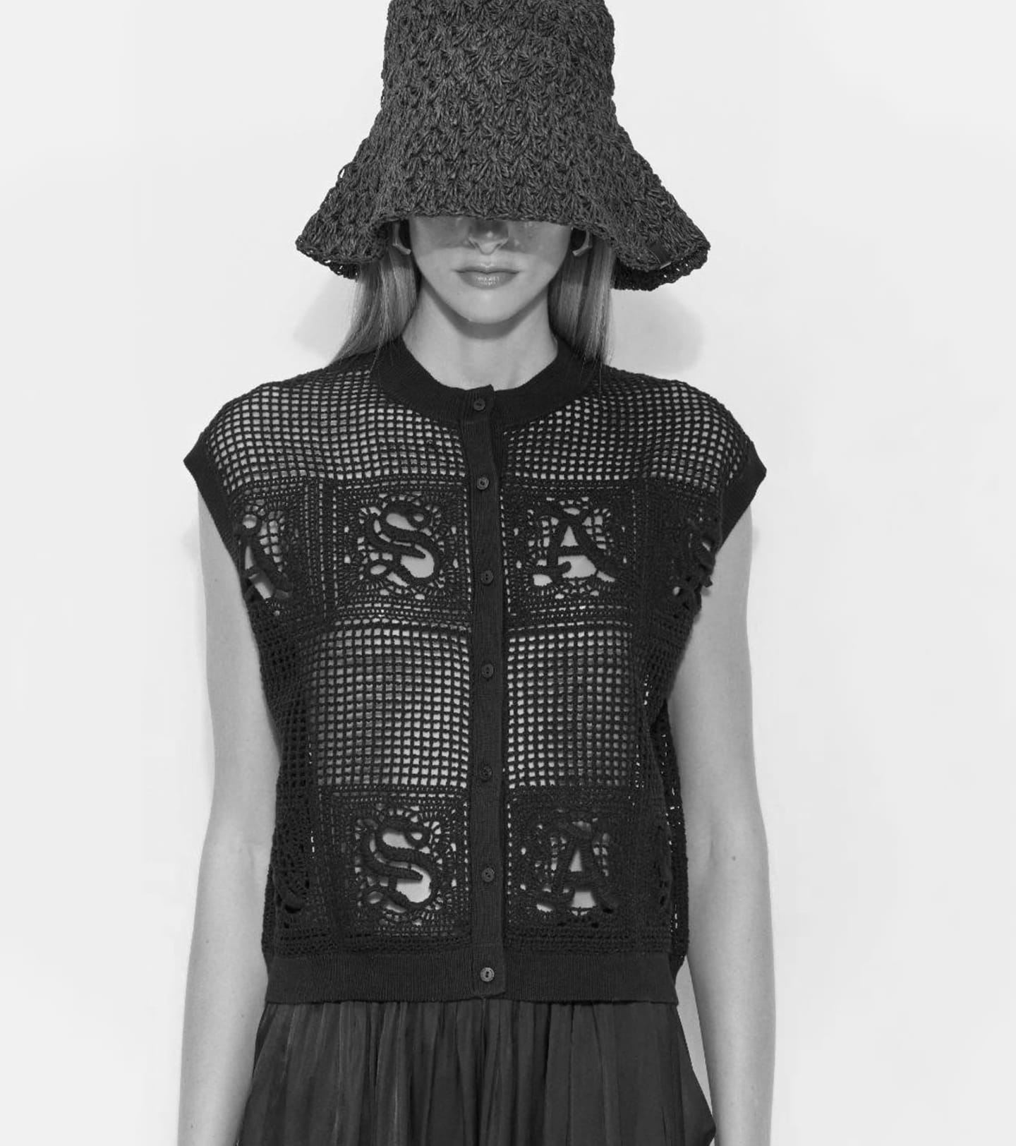 A fashion model wearing a dark, wide-brimmed crochet bucket hat and a matching sleeveless, open-knit black button-up vest.