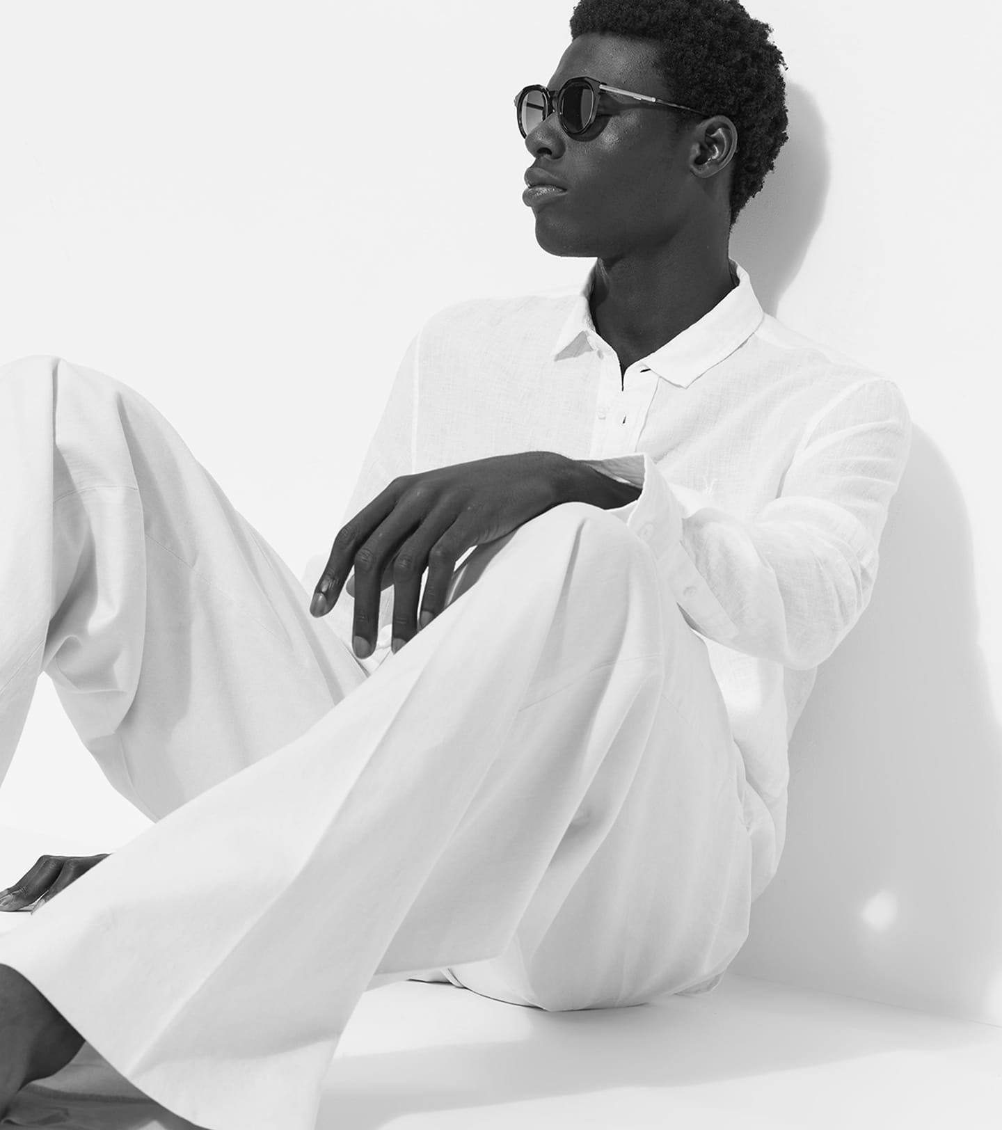 A man wearing a long-sleeved white polo shirt and wide-leg white trousers, sitting against a white wall while wearing dark sunglasses, presented in a black-and-white editorial style.
