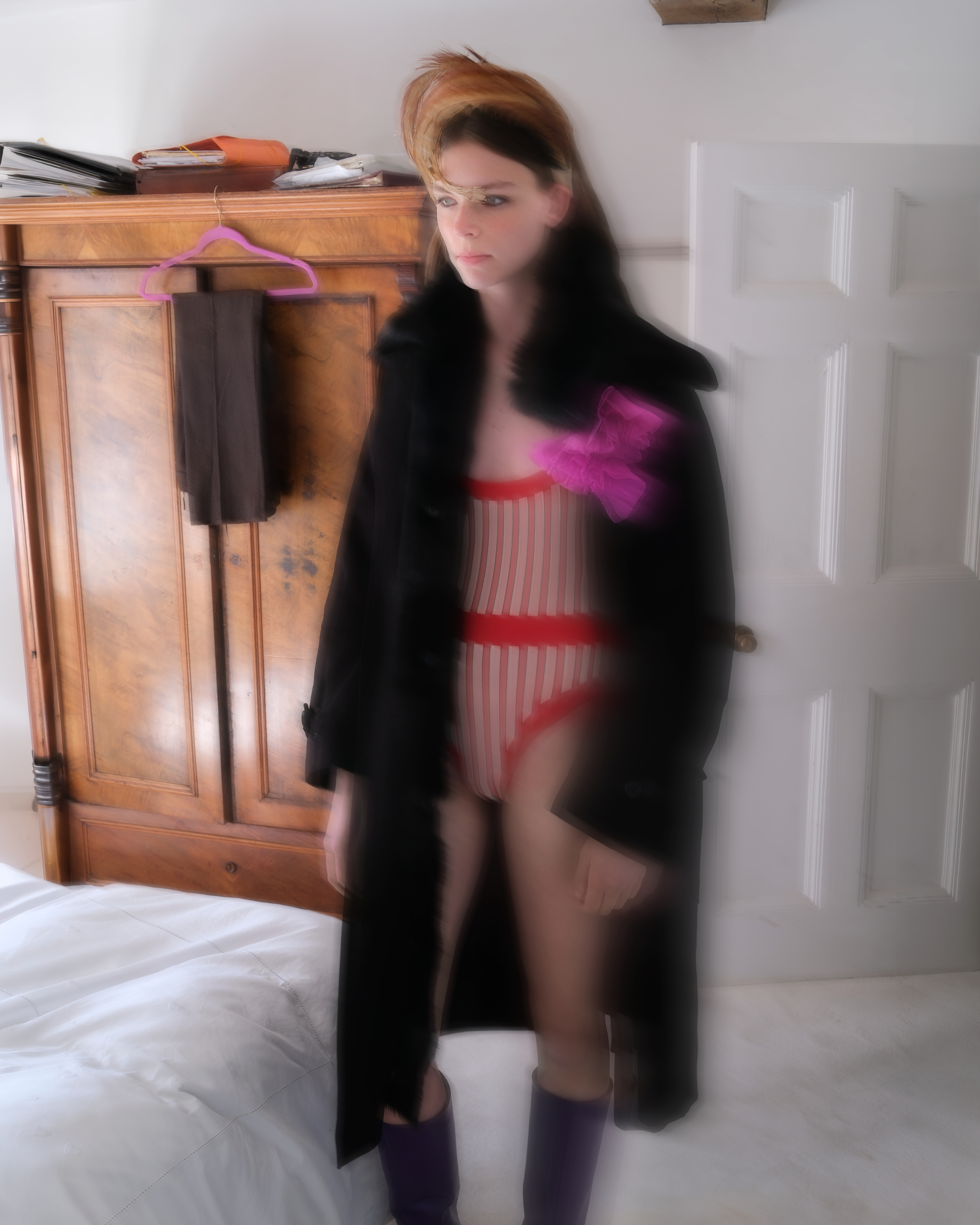 Model standing in a bedroom wearing a black coat over a red-and-white outfit, with a wooden wardrobe behind her.