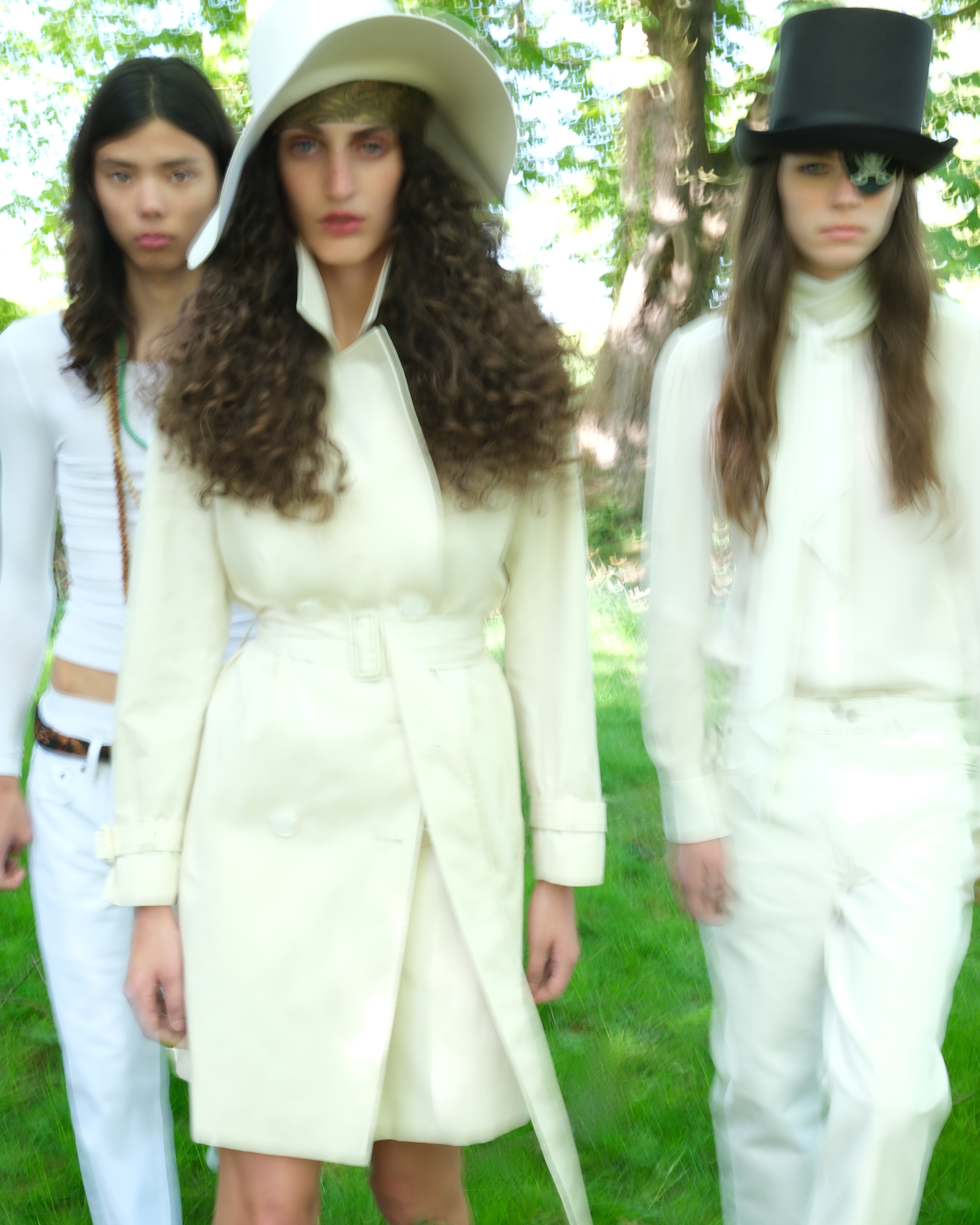 Three models wearing white outfits and hats standing together outdoors on grass.