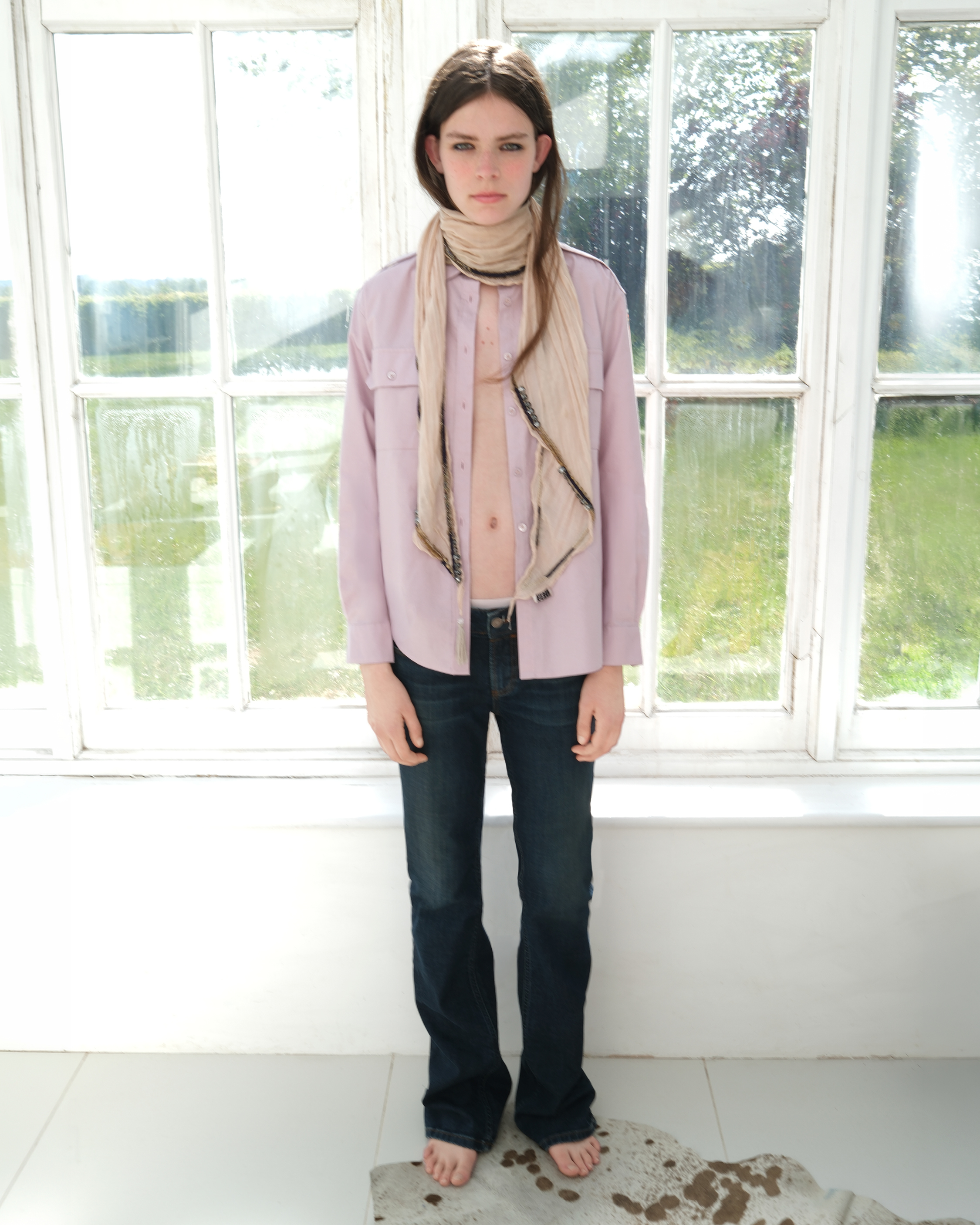Model wearing a pale pink jacket and scarf, standing indoors in front of large windows.