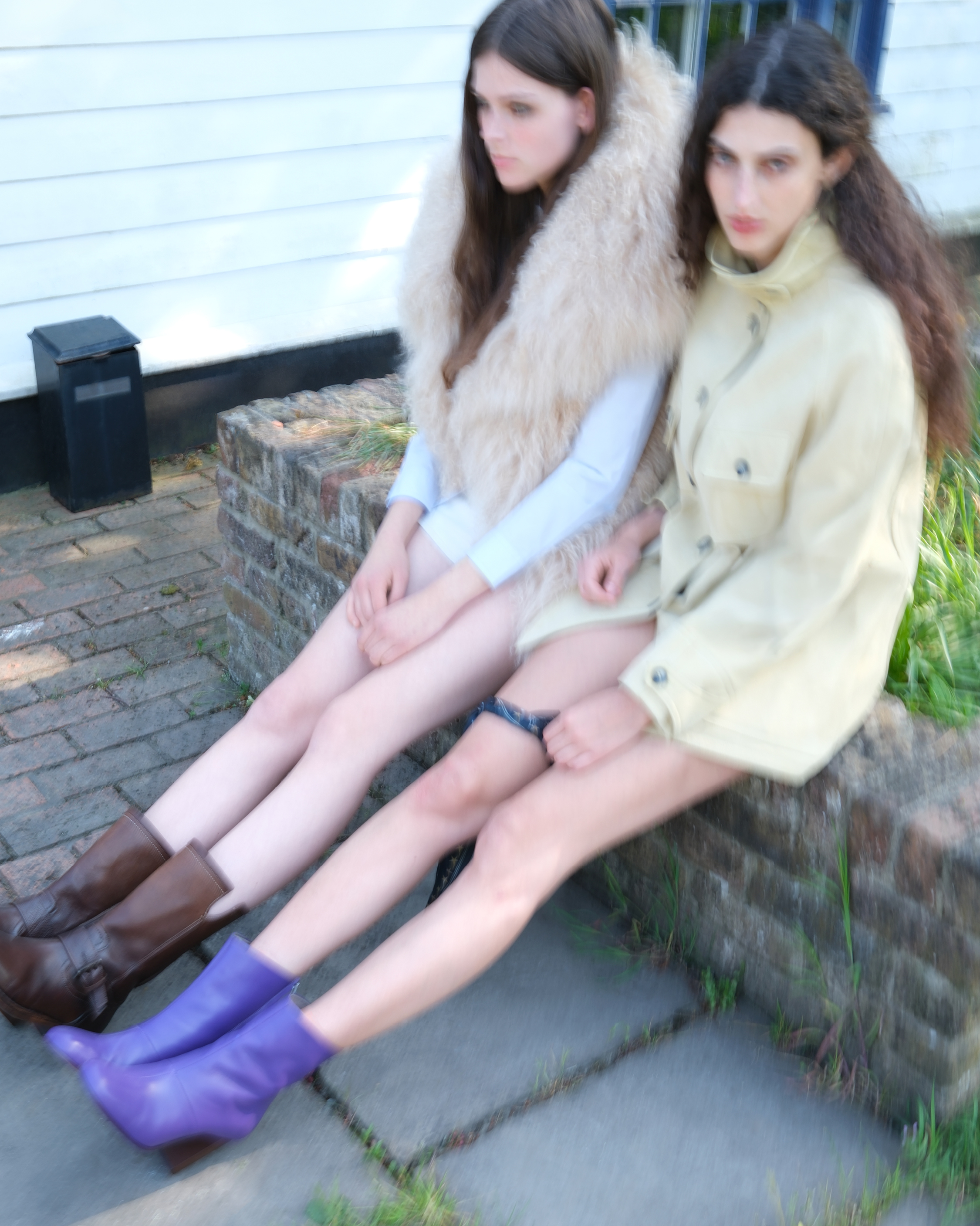 Two models seated outdoors on a low brick wall wearing layered neutral-toned outfits and boots.