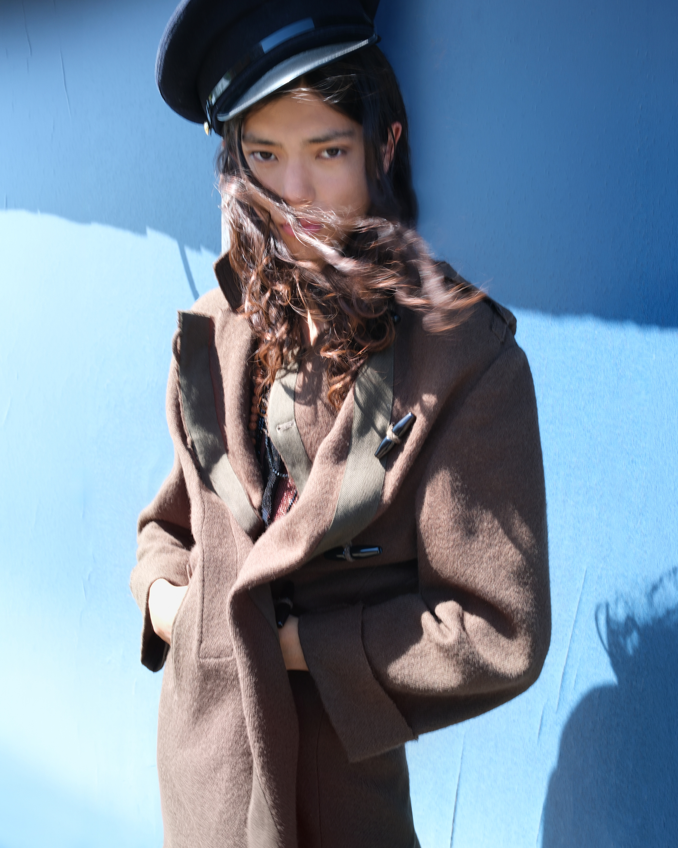 Model wearing a brown blazer and black peaked cap, photographed against a blue wall.