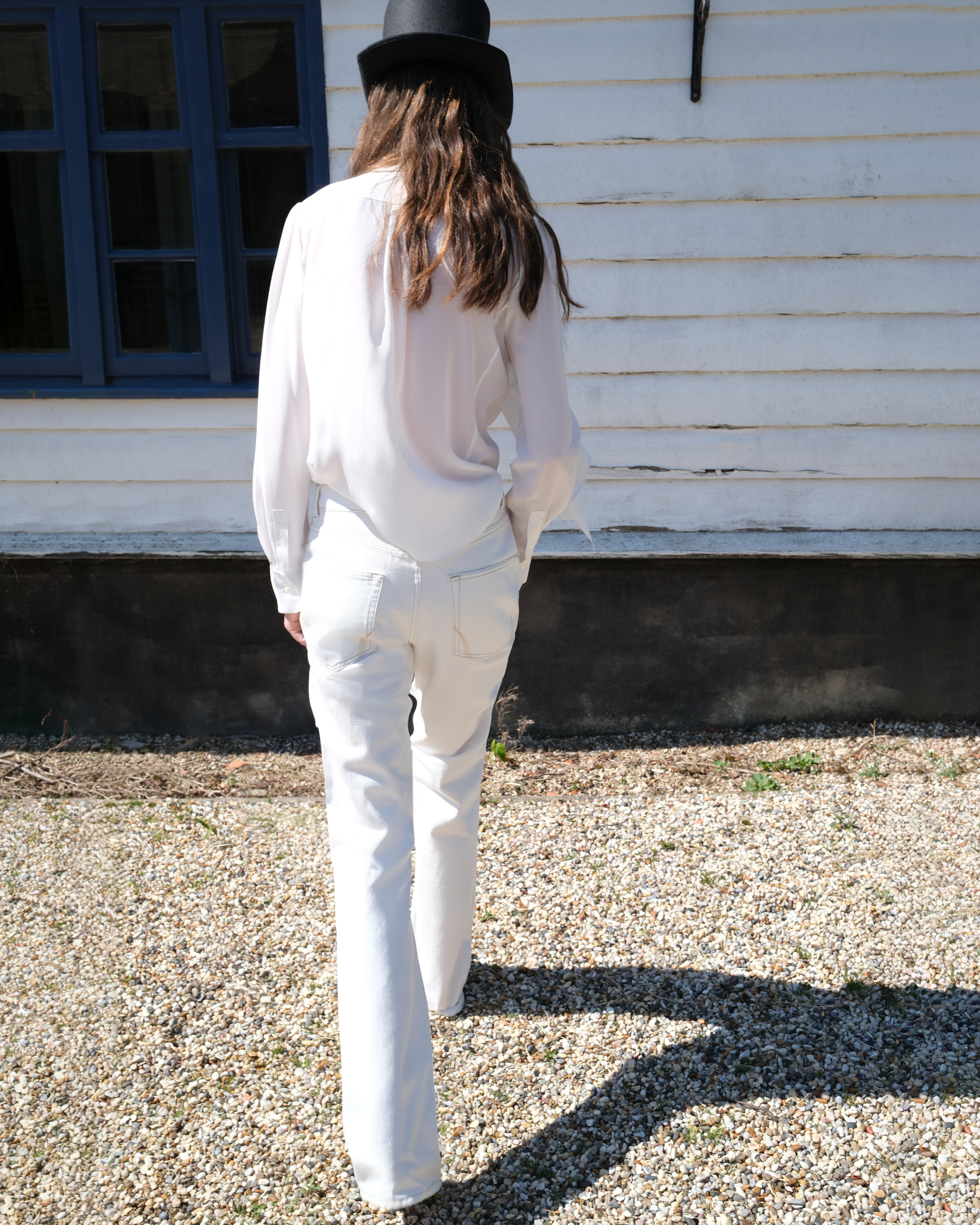 Person dressed in white walking outdoors beside a white wooden building, photographed from behind.