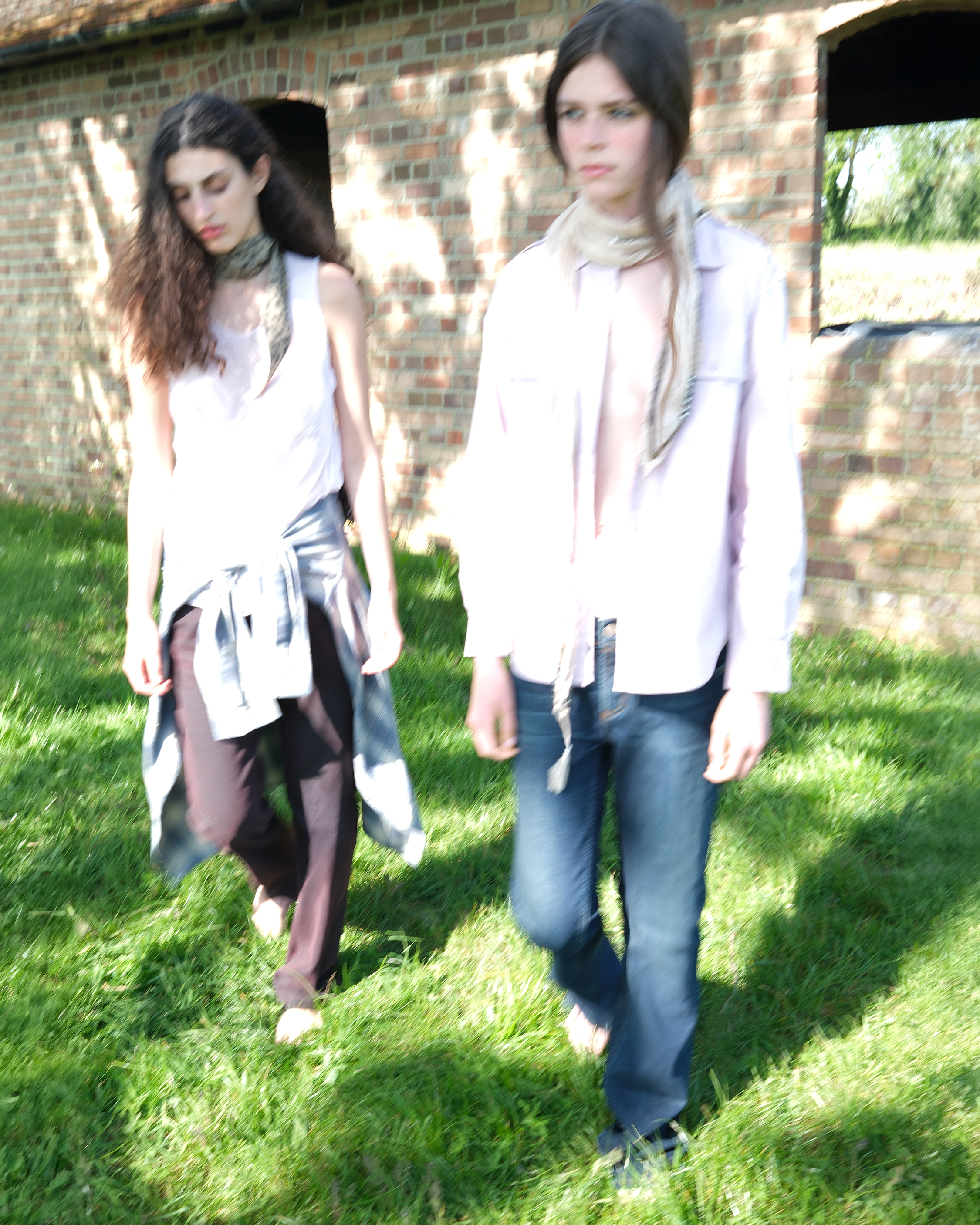 Two models walking outdoors on grass in layered light-toned outfits.