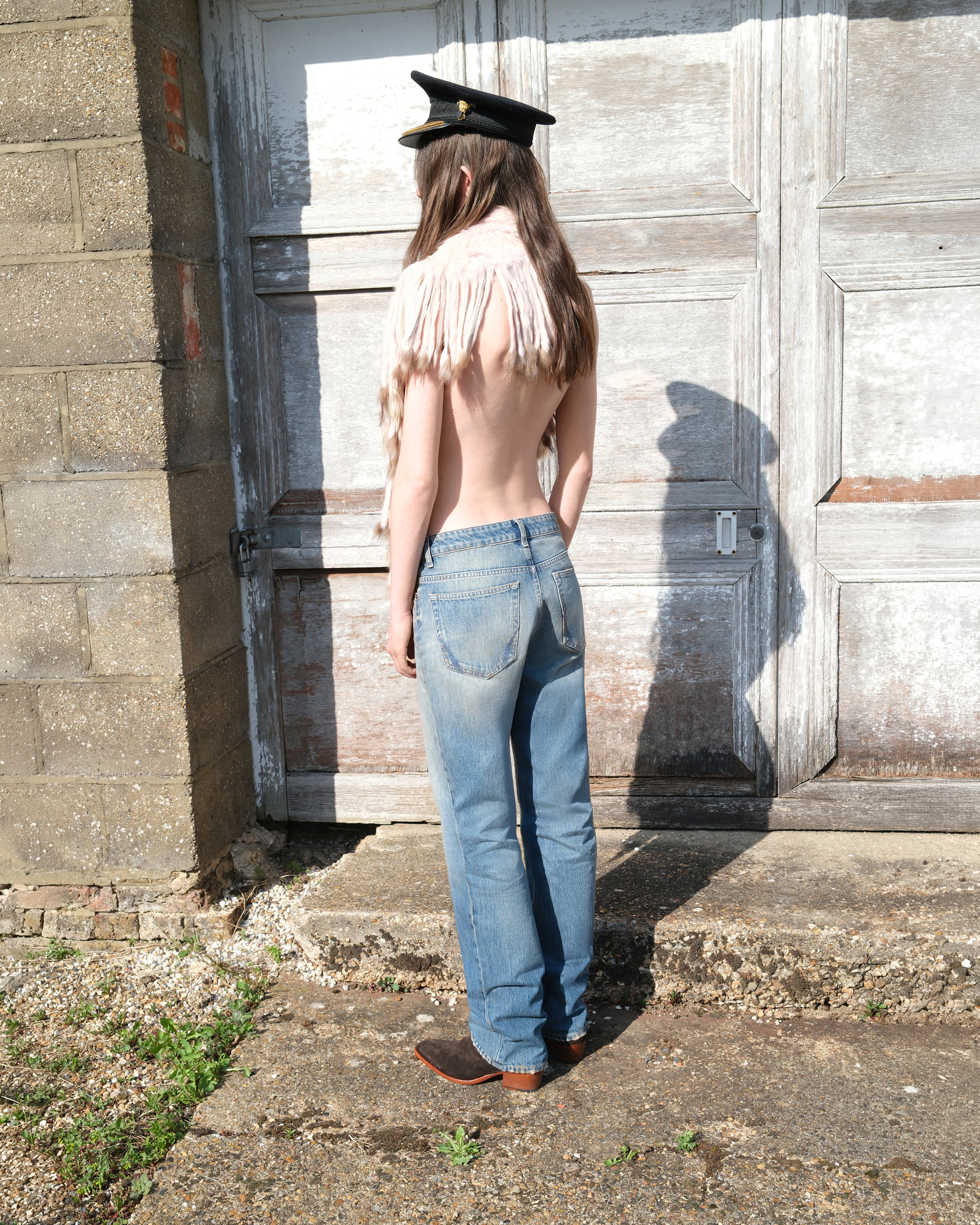 Model wearing jeans and a black peaked cap, photographed from behind outside a weathered wooden building.