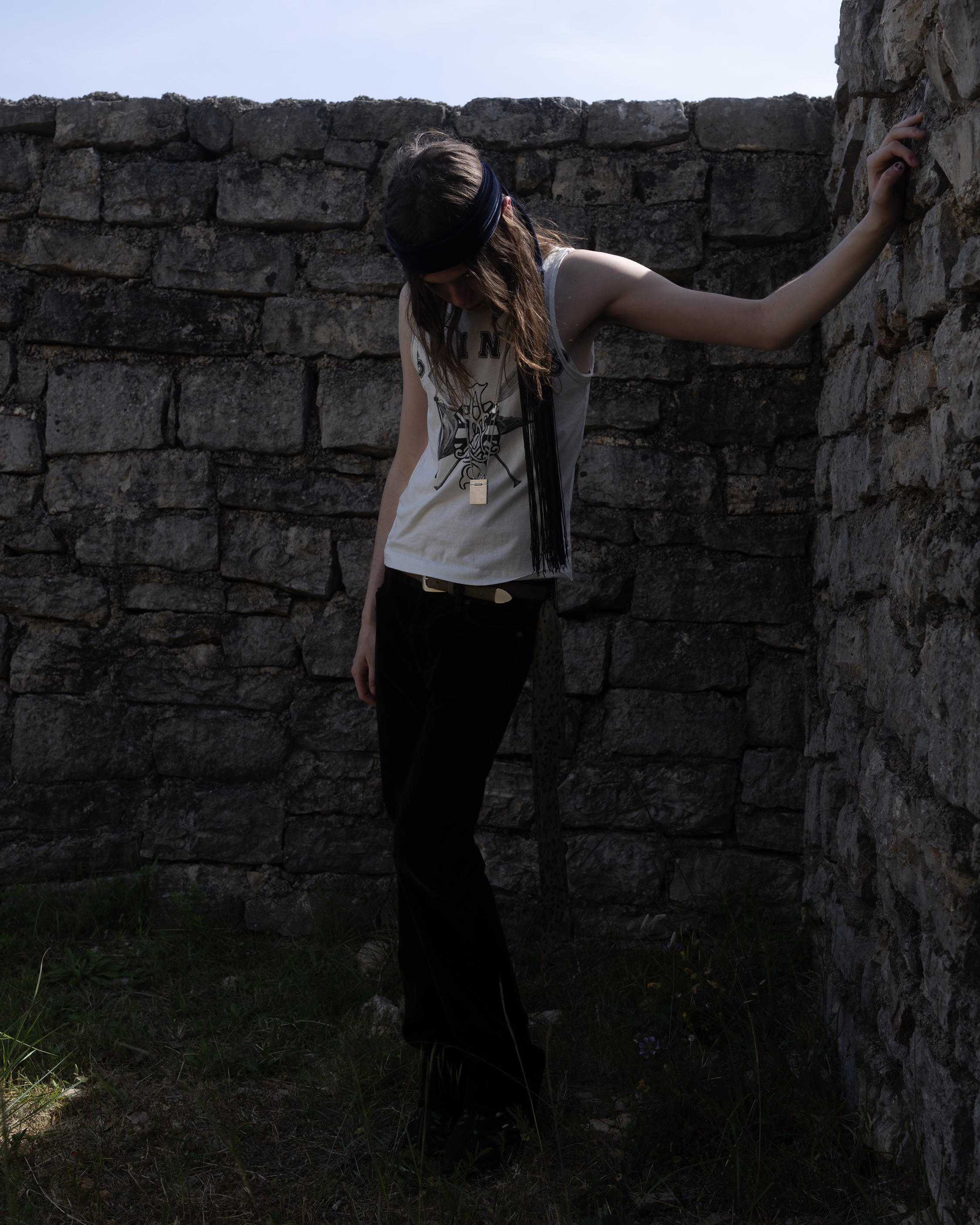 Portrait of a person standing beside a stone wall outdoors, wearing a sleeveless top and black trousers.