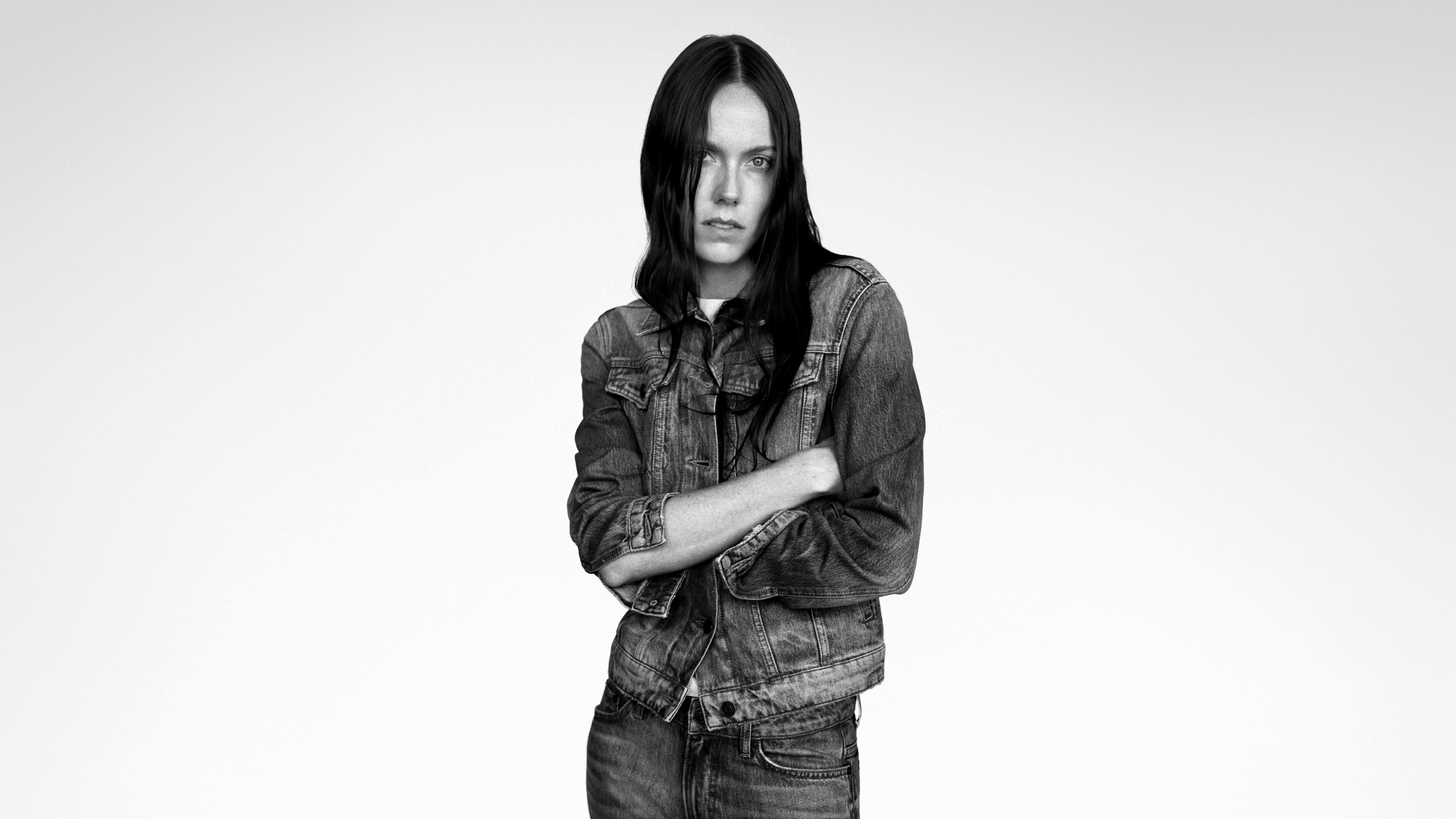 A black and white portrait of a woman with long dark hair wearing a denim jacket and jeans, standing with her arms crossed against a pale grey background.