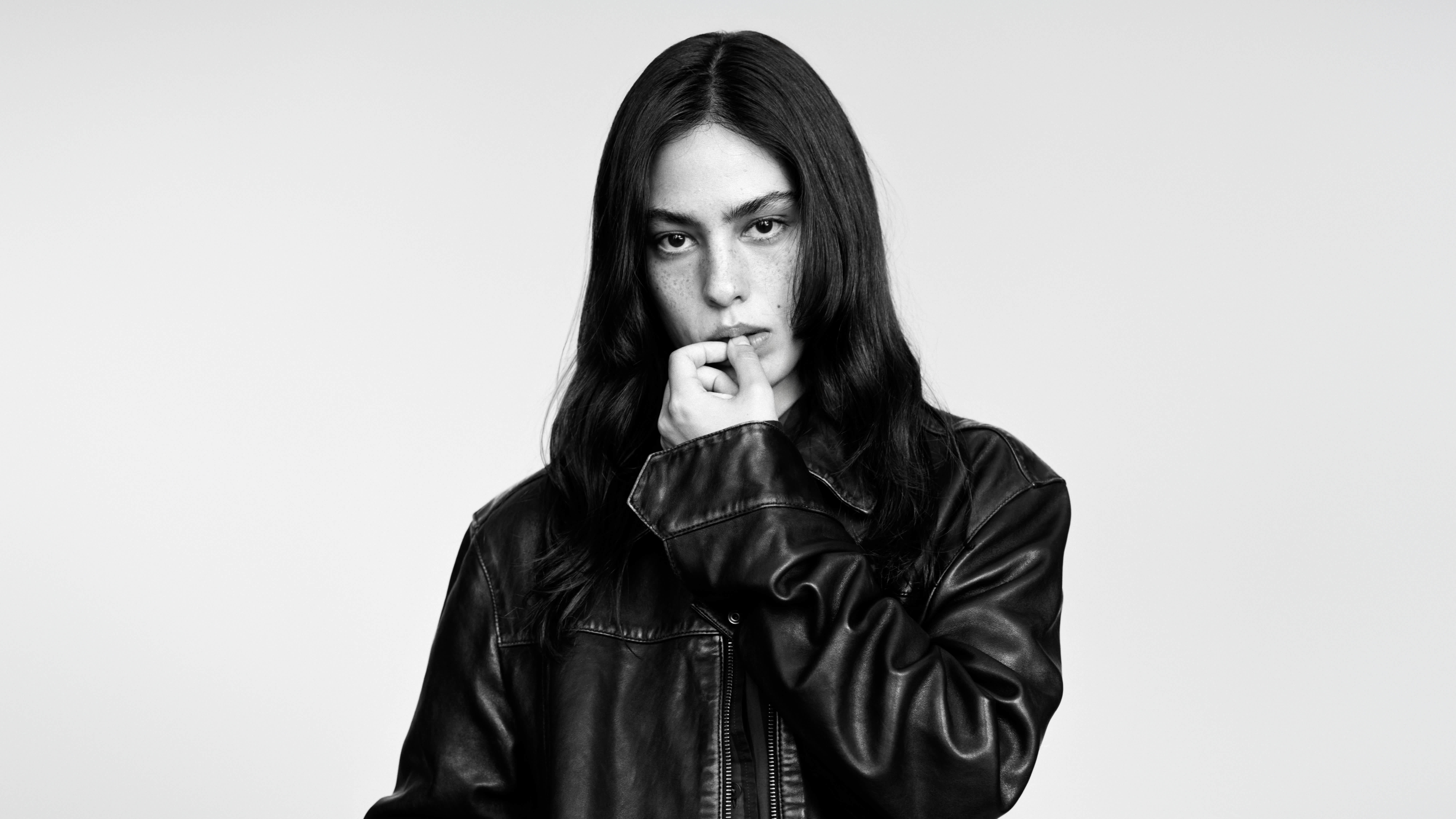 A black and white portrait of a woman with long dark hair posing in a black leather jacket, set against a plain light grey background.