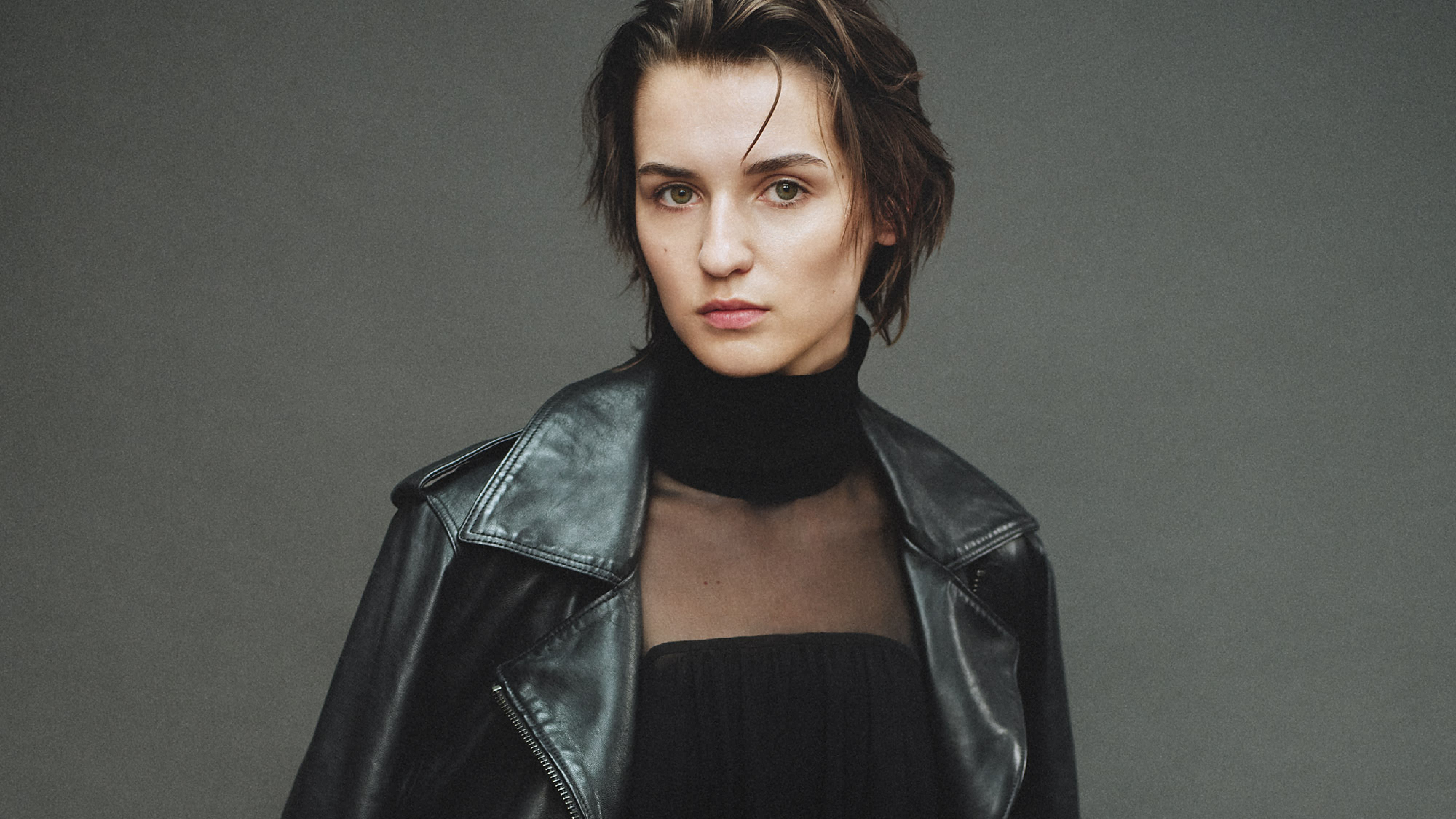 A woman with short brown hair wears a black leather jacket and sheer black top, posing in front of a neutral grey background
