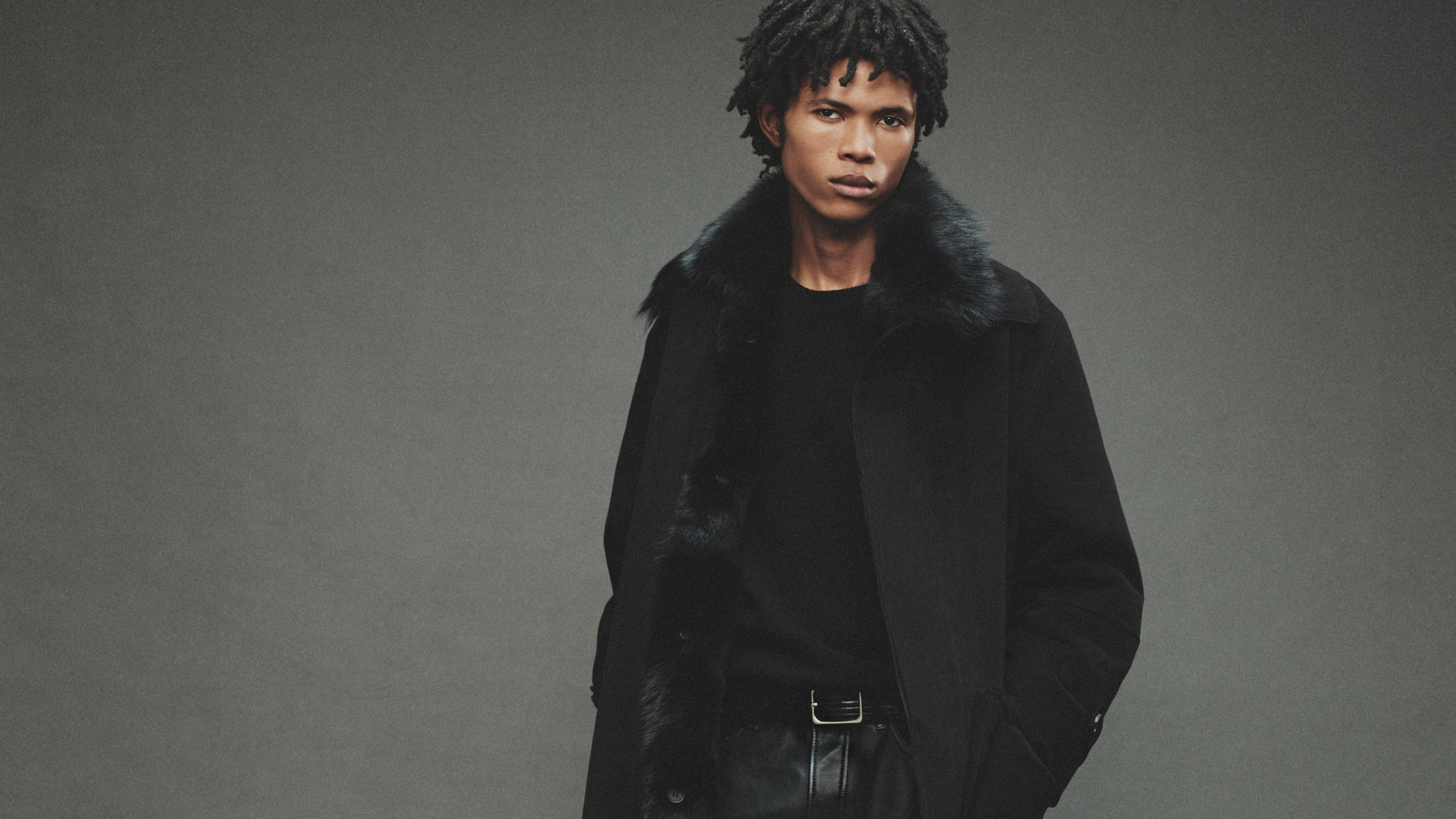 A man with textured hair wears a black coat with a fur collar over a black shirt, posing in front of a neutral grey background
