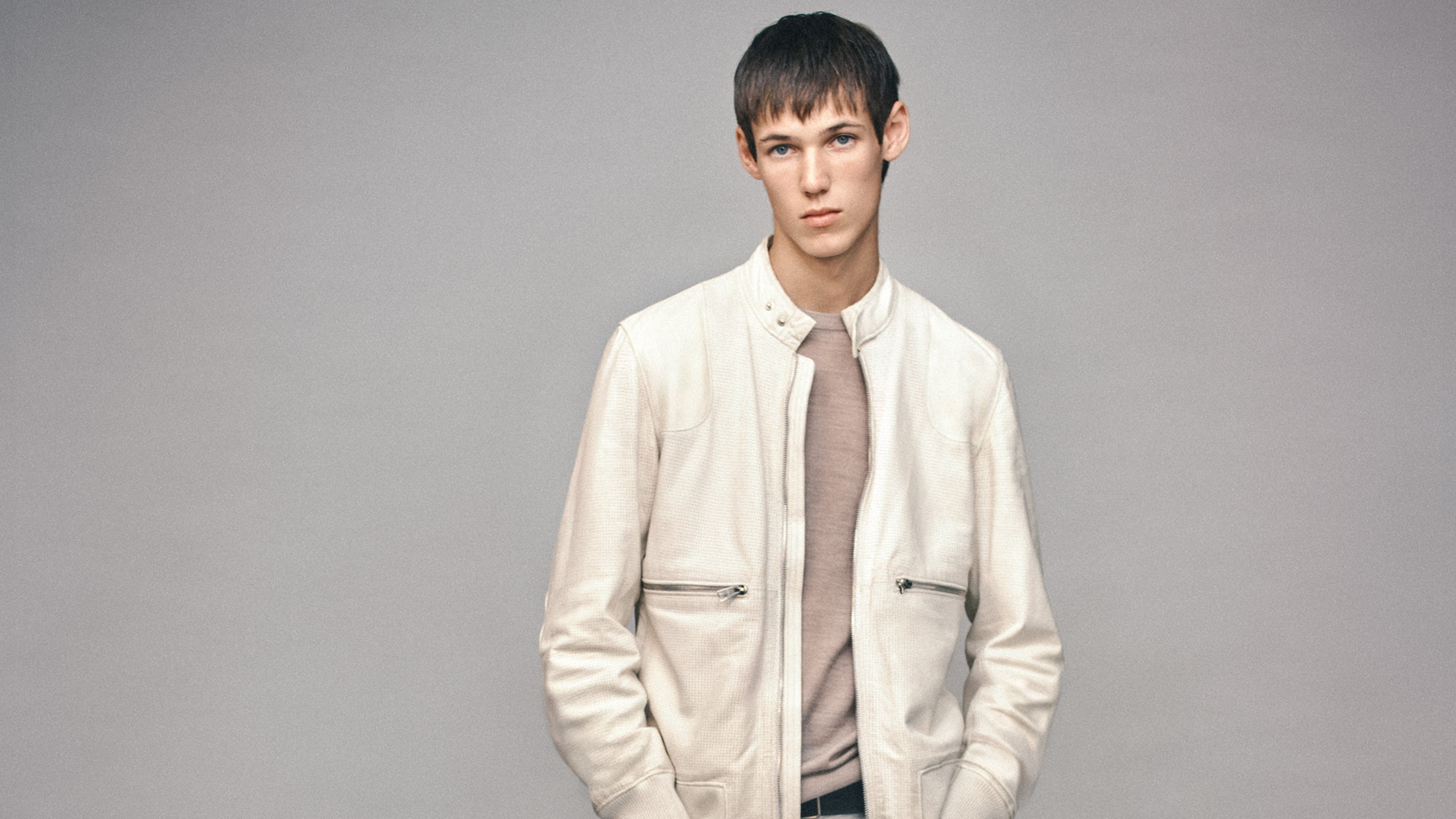 A man with short brown hair wearing a cream leather jacket and beige shirt stands against a grey background.