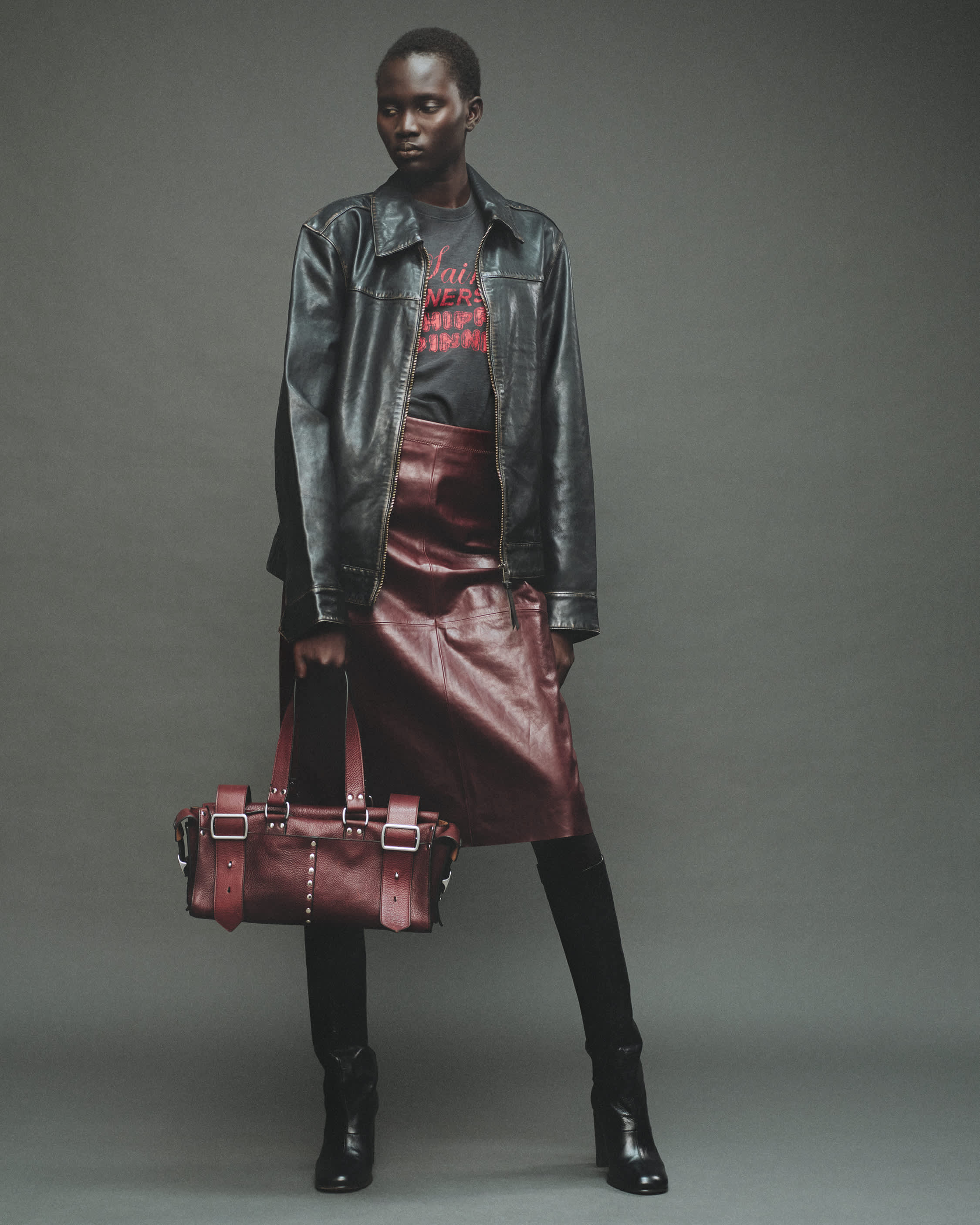 A woman with short black hair stands against a grey background wearing a black leather jacket, a oxblood red leather skirt, and holding a oxblood red leather handbag.
