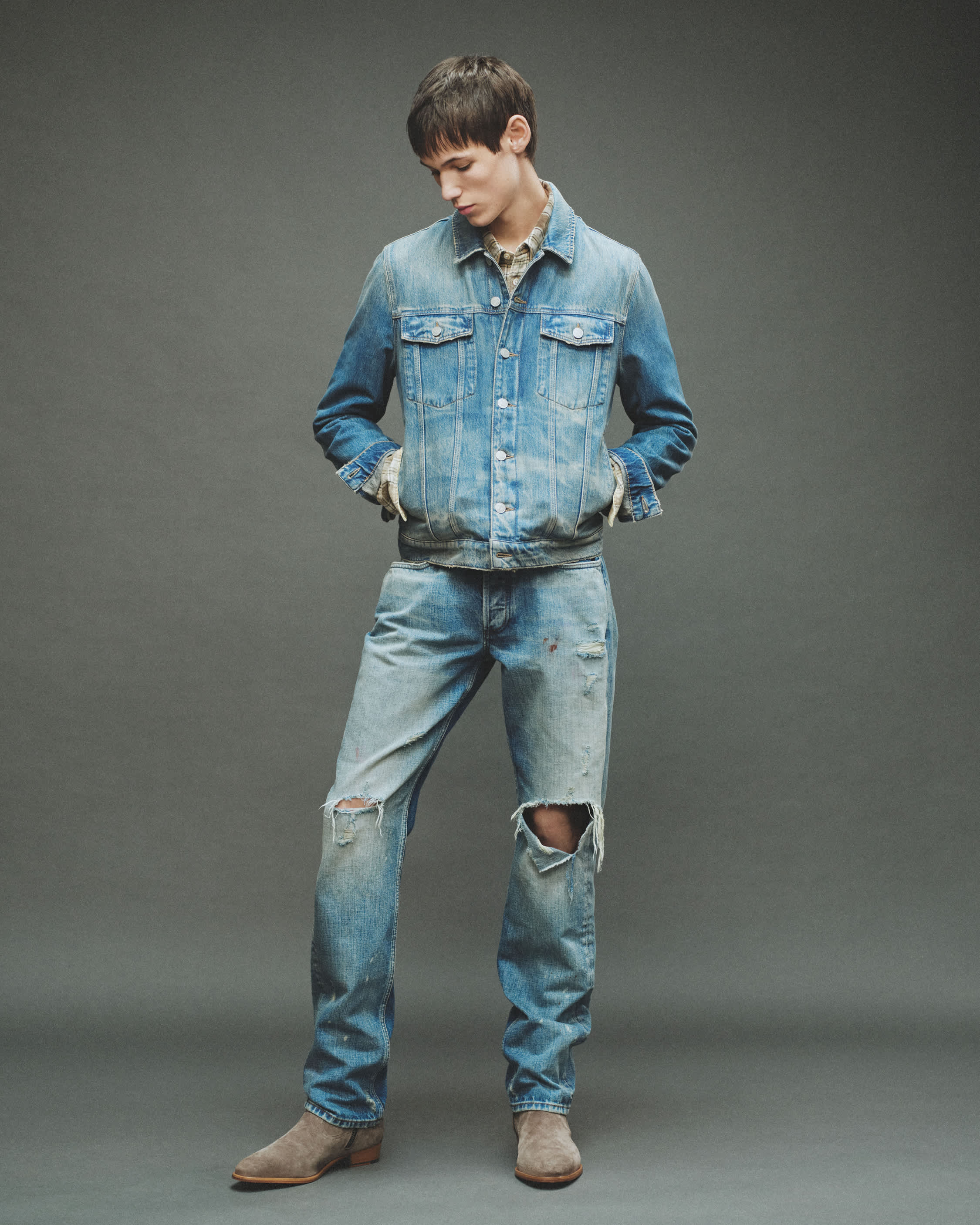 A man with short brown hair looking down to the right, wearing a light blue denim jacket and matching distressed jeans with knee rips, standing against a neutral grey background