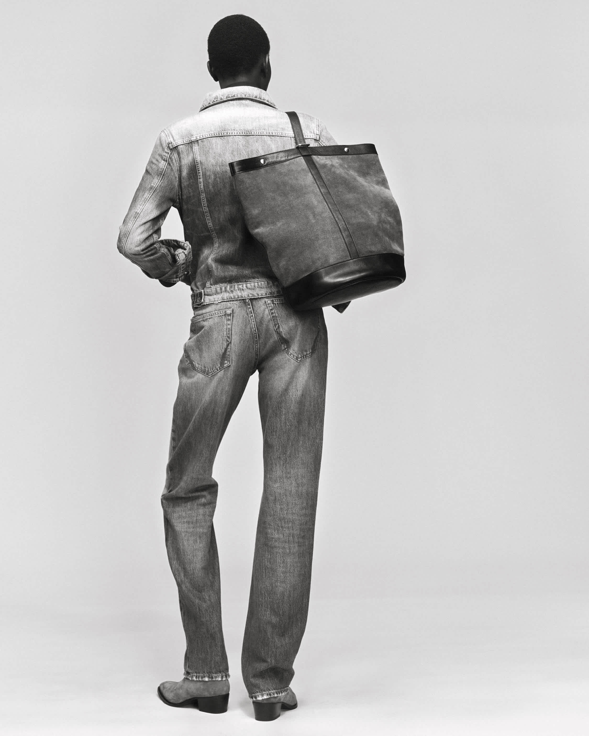 A woman with short black hair wearing jeans and a denim jacket carrying a bag over the shoulder, stands upright, with her back towards the camera.