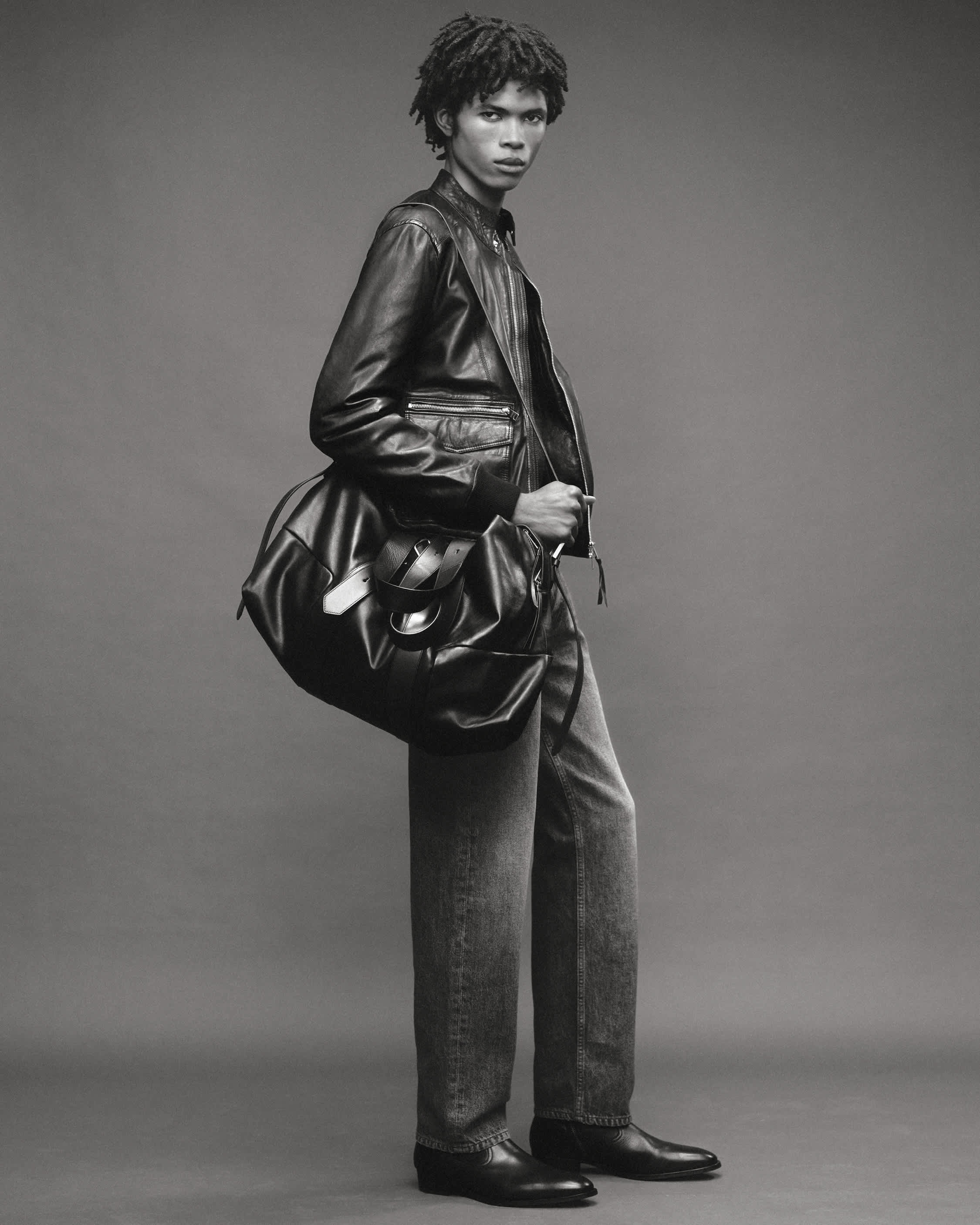 A man with short textured hair wears a black leather jacket and carries a large black leather bag, standing against a plain grey background.